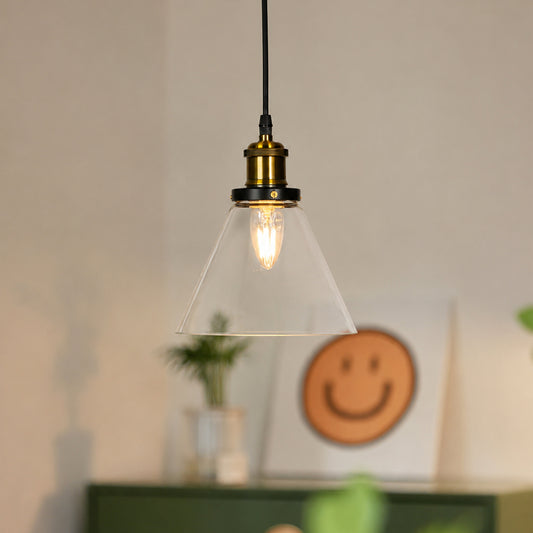 A hanging pendant light with a clear glass cone shade, brass metal socket, and black cord, shown lit in a softly blurred indoor setting.