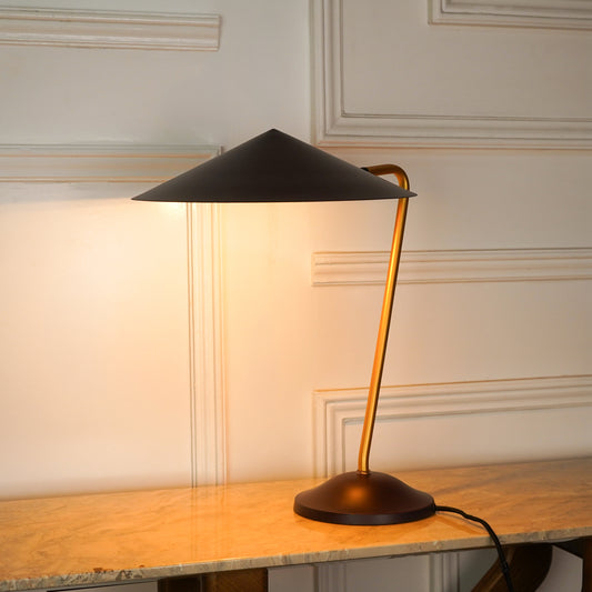 A modern table lamp with a black conical shade, a curved gold-toned metal stem, and a round dark base, illuminated and placed on a light wood console table against an off-white paneled wall.