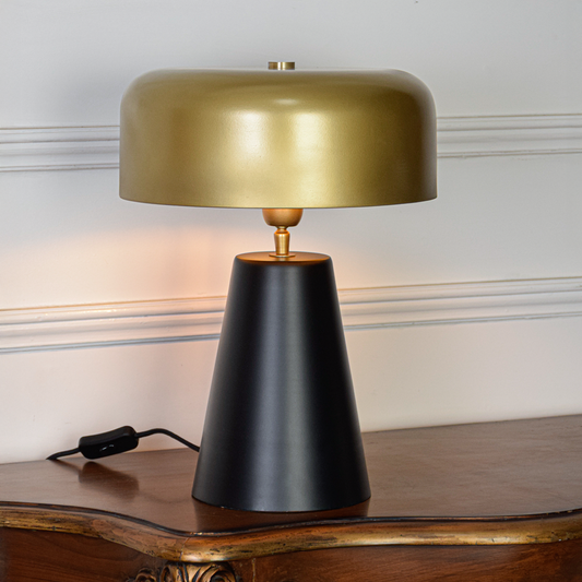 Table lamp shown on white background with gold dome shade black tapered base and soft glow highlighting modern form and premium finish.