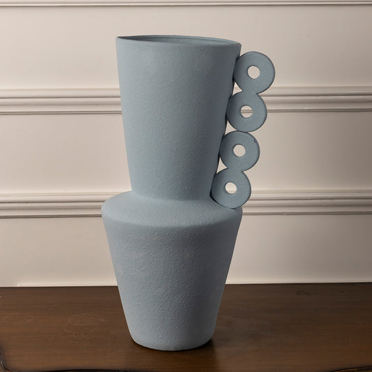 Pastel blue ceramic vase with stacked form and circular handle accents placed on wooden table against neutral wall background for contemporary decor appeal.