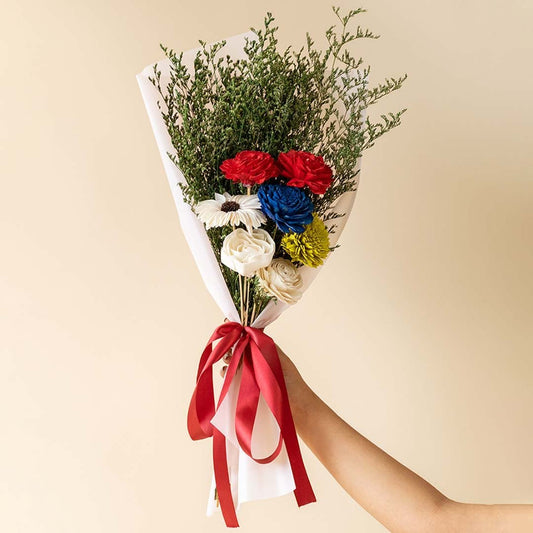 Main lifestyle image showing preserved multicolor sola flower bouquet with green limonium grass wrapped in white paper and red ribbon held by hand in elegant setting.