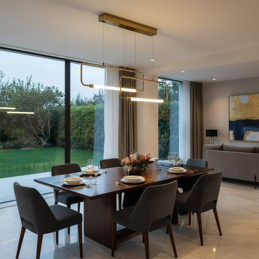 Gold linear pendant light hanging above wooden dining table with set place settings beside large windows creating elegant modern dining space.