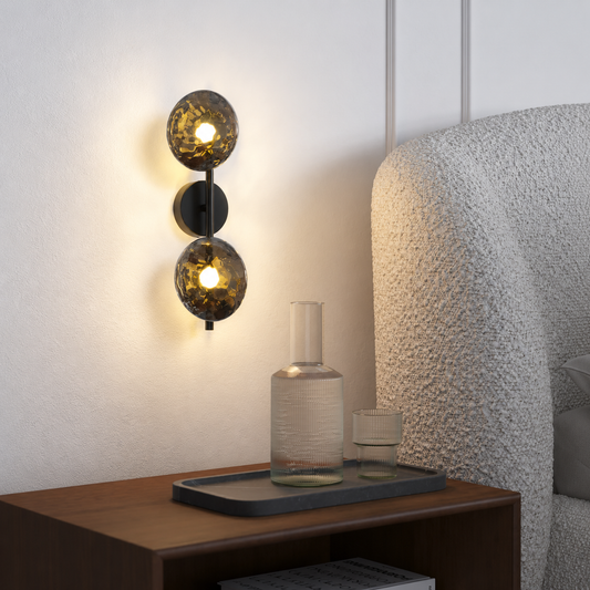 A modern wall-mounted sconce with two spherical amber-tinted glass shades and a black metal body, shown lit on a bedroom wall above a wooden bedside table.