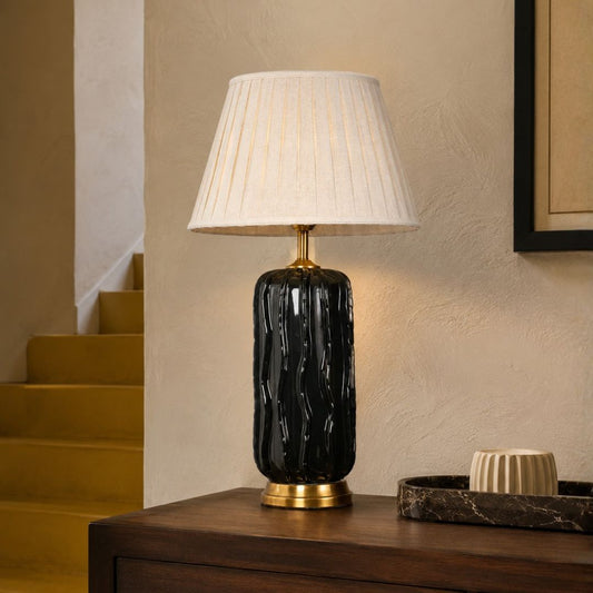 A table lamp with a black textured cylindrical base and a pleated off-white shade, sitting on a dark wooden table in a warm-toned interior setting.