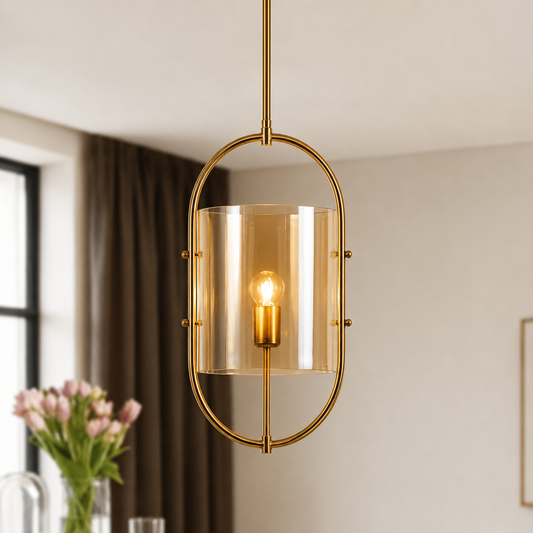 A modern brass pendant light with a clear amber-tinted glass cylinder shade and a visible warm Edison bulb, hanging in a softly lit interior room.