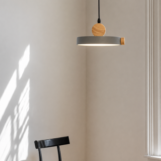 A modern pendant light with a matte gray circular shade, a round natural wood accent, and a black cord, hanging in a minimalist room corner with warm sunlight and window shadows.