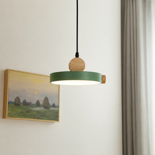 A modern pendant light with a matte green circular shade, natural wood finials, and a black cord, hanging in a minimalist room next to a window and a framed landscape painting.