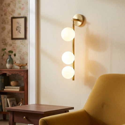 A modern wall-mounted light fixture with three vertical frosted glass globes on a slim brushed brass bar, shown lit in a warm interior setting beside a mustard chair and wooden side table.