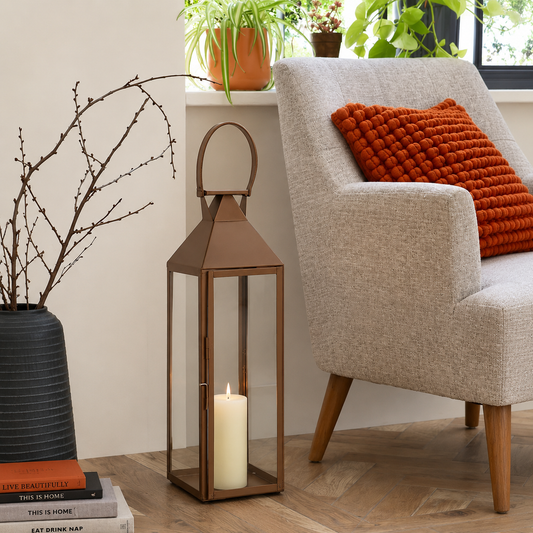 Rustic bronze metal tall frame glass lantern floor candle holder next to an armchair for cozy living room home decor styling.