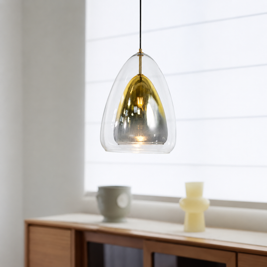 A modern pendant light with a clear teardrop-shaped glass shade and a brushed gold interior, hanging in a bright, minimalist room with a wooden sideboard and white window blinds.
