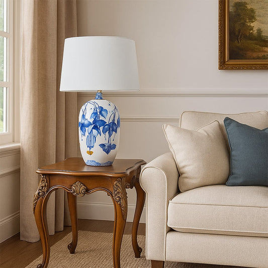 White ceramic table lamp with blue floral artwork and a soft white shade placed on a carved wooden side table adding a calm elegant touch to living room decor.