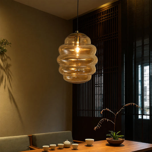 A modern pendant light with a ribbed, layered clear glass shade hanging over a wooden dining table in a warm, dimly lit interior.