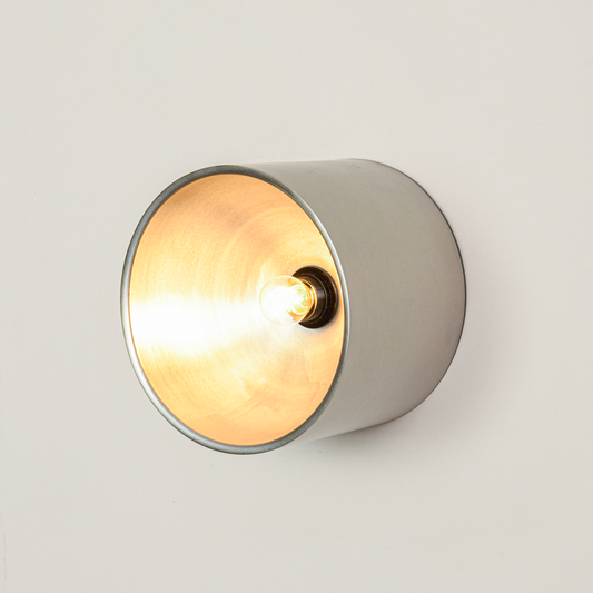 Wall mounted circular metal light switched on against clean white background showing warm golden inner glow and minimalist cylindrical outer body.