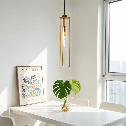 A modern brass pendant light with a clear glass cylinder hangs over a white dining table in a bright room. A framed Matisse print and a green monstera plant in a glass vase sit on the table, with a large window on the right.
