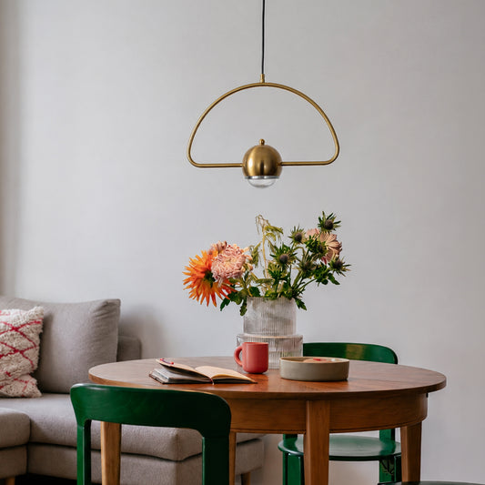 A modern brass pendant light with a semi-circular frame and a dome-shaped bulb holder hangs over a round wooden dining table in a bright, contemporary room. The table is styled with a vase of colorful flowers, a red mug, and books, surrounded by green chairs.