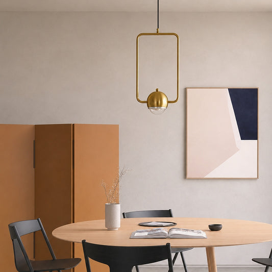 A modern dining room scene featuring a rectangular gold pendant light hanging above a light wood table with black chairs. The background includes a warm beige wall, a geometric abstract art print, and a terracotta-colored cabinet.
