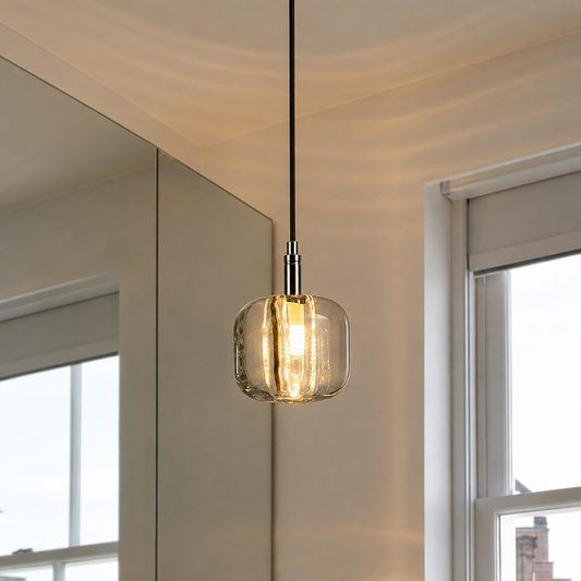 A modern pendant light with a clear glass cylindrical shade and a black cord, hanging in a room with a window and mirror visible.