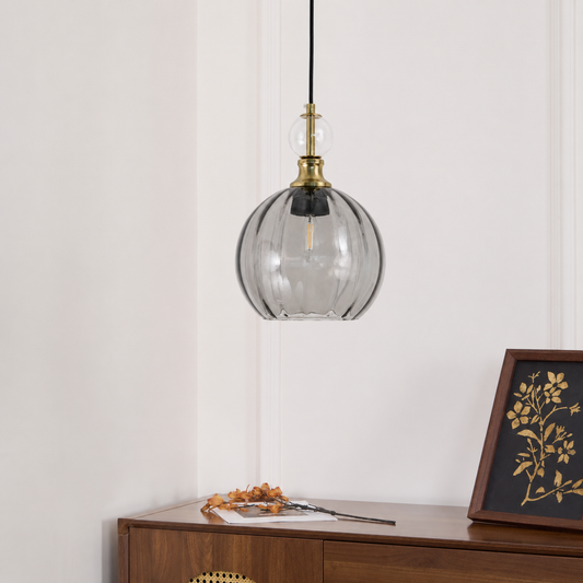 A modern pendant light with a ribbed clear glass globe shade and a brass-finished top cap, hanging above a wooden console table in a styled interior setting.