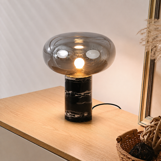A modern table lamp with a rounded, dome-shaped smoky glass shade and a black marble base, illuminated on a light wood tabletop.