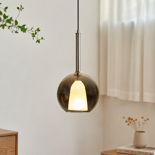 A modern pendant light with a dark bronze glass globe shade and a warm glowing frosted center, hanging in a softly lit room with curtains, a small wooden table, and dried flowers in the background.