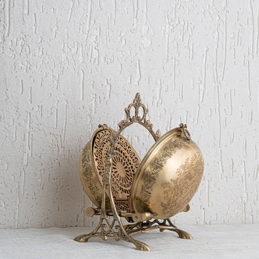 A brass swing bowl with floral carving is displayed on a plain white background showing its hinged lid delicate frame and intricate openwork back plate.