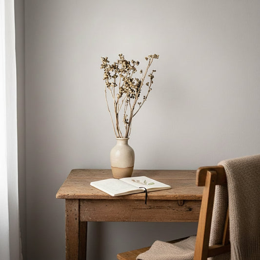 A rustic natural beige dried button flower bouquet in a ceramic vase resting on a wooden study desk by a window setup.