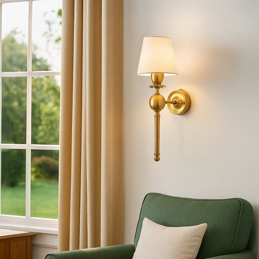 Classic gold torch wall sconce lamp with a cream fabric shade casting warm light over a cozy green living room armchair.