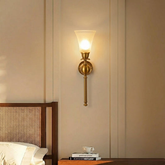 A warm brass wall sconce with a frosted glass shade glowing with a soft yellow light, mounted on a beige paneled wall above a wooden headboard and bedside table.
