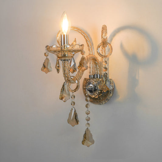 A decorative wall sconce with a lit bulb, featuring a warm gold-toned metal frame, clear faceted glass crystal drops, and a curved arm, mounted on a plain light wall with a soft shadow cast to the right.
