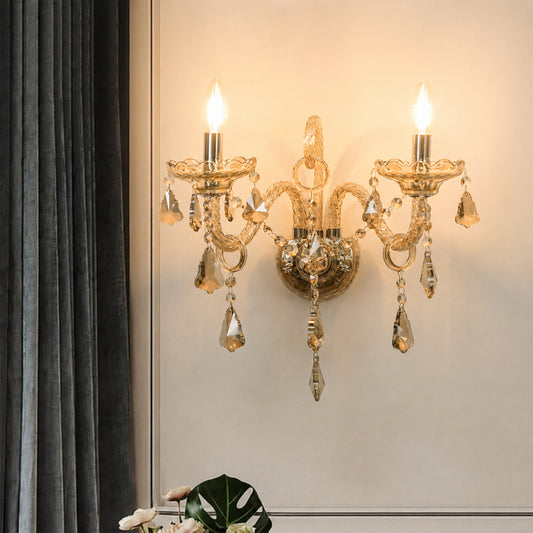 An ornate wall-mounted sconce with two candle-style bulbs, a gold-toned metal frame, and hanging clear crystal droplets, shown lit on a beige wall next to dark gray curtains.