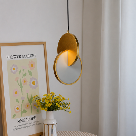 A modern pendant light with overlapping circular gold and white shades hanging in a bright room next to a framed 'Flower Market Singapore' print and a vase of yellow flowers.