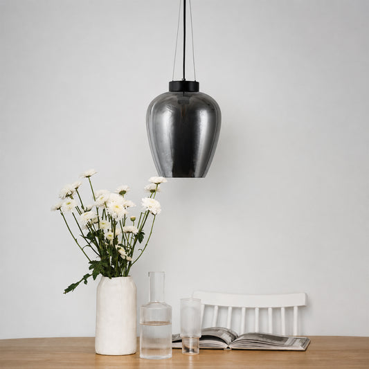 A modern teardrop-shaped pendant light with a brushed metallic silver finish hangs over a wooden table styled with a white vase of daisies, a glass carafe, a glass, and an open magazine against a plain light gray wall.