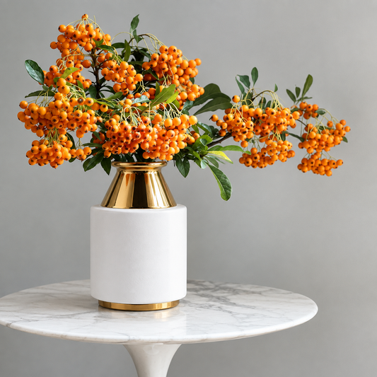 A white cylindrical vase with a metallic gold geometric neck and base, filled with bright orange berries and green leaves, displayed on a white marble round table against a neutral gray background.