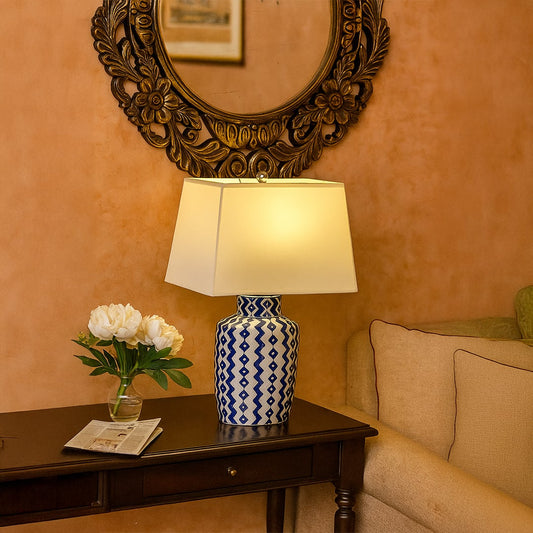 Blue patterned lamp with yellow shade on side table with light on and tan wall with carved mirror background for vibrant décor.