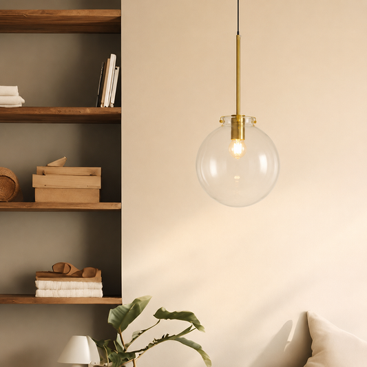 A modern pendant light with a clear glass globe shade and a brushed brass rod hanging in a minimalist room setting.