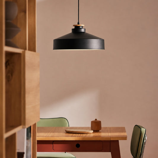 A matte black pendant light with a warm wood accent ring hangs over a wooden dining table in a minimalist room with beige walls.