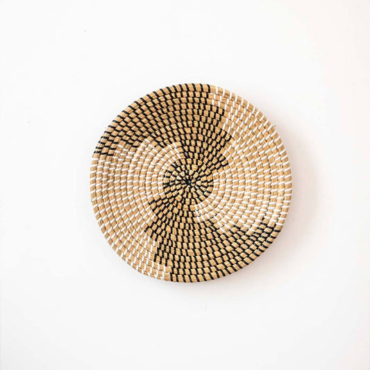 Abstract spiral patterned black and beige woven seagrass wall hanging basket plate isolated on a white backdrop.