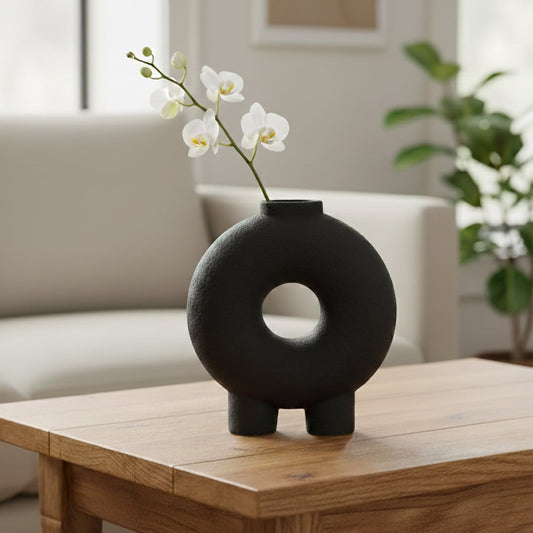 Black textured ceramic decorative ring vase styled with orchid stem on wooden coffee table in bright modern living room.