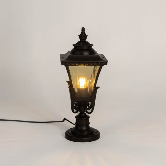 Designer outdoor black metal pillar lamp glowing with warm amber glass shade perfect for patio terrace and driveway.