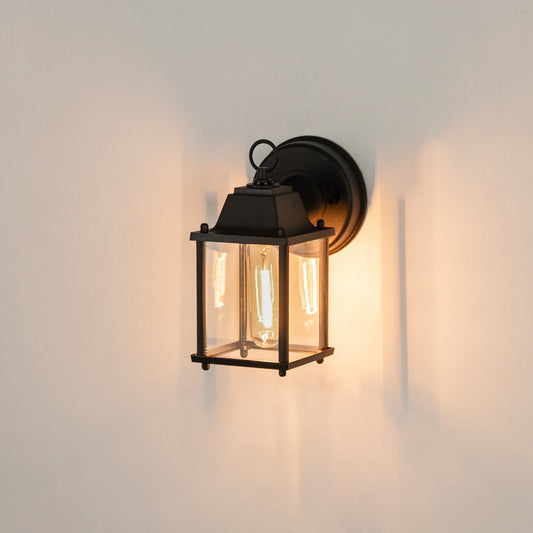 Modern black rectangular outdoor wall light on white background with clear glass panels casting a warm downlight glow.