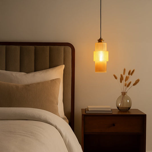 Warm glowing pendant light with ribbed glass and brass finish hanging beside bed for modern bedroom lighting.