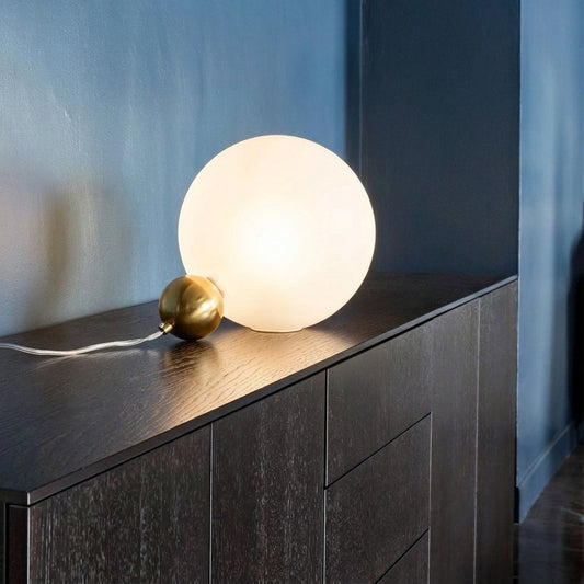 Stylish glowing globe table lamp with brass metal base placed on dark console against blue wall background perfect for cozy ambient lighting.