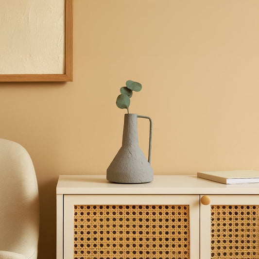 Gray textured ceramic jug with handle holding fresh eucalyptus stems displayed on wooden desk with rattan cabinet in scandinavian home office decor.