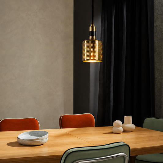 A modern cylindrical pendant light with a textured gold mesh shade hangs over a wooden dining table in a styled interior setting with chairs and neutral decor.