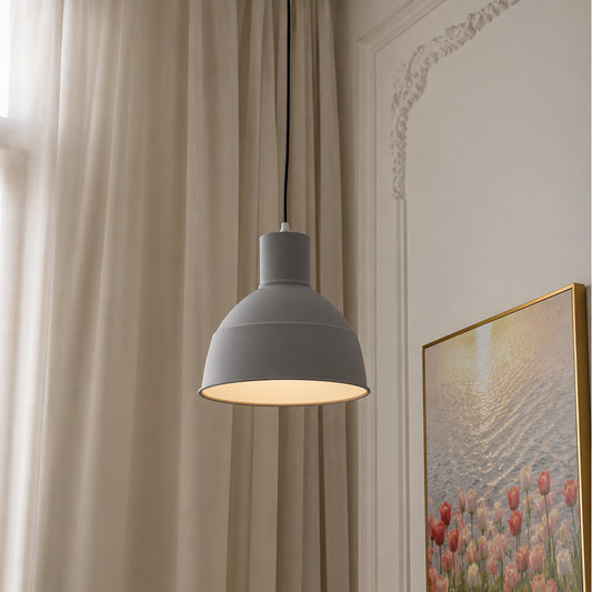 A modern gray pendant light with a matte finish and a warm glowing interior, hanging in a styled room next to beige curtains and a framed floral print.