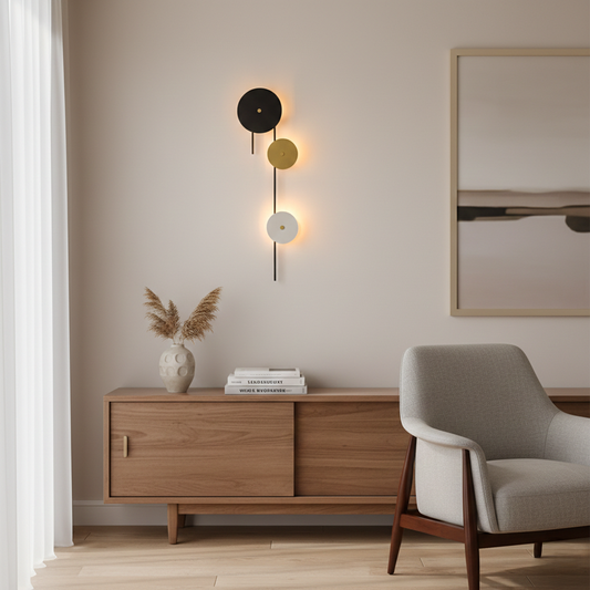 Modern wall light with layered black gold and white discs glowing softly above wooden sideboard beside lounge chair in warm minimalist living room.