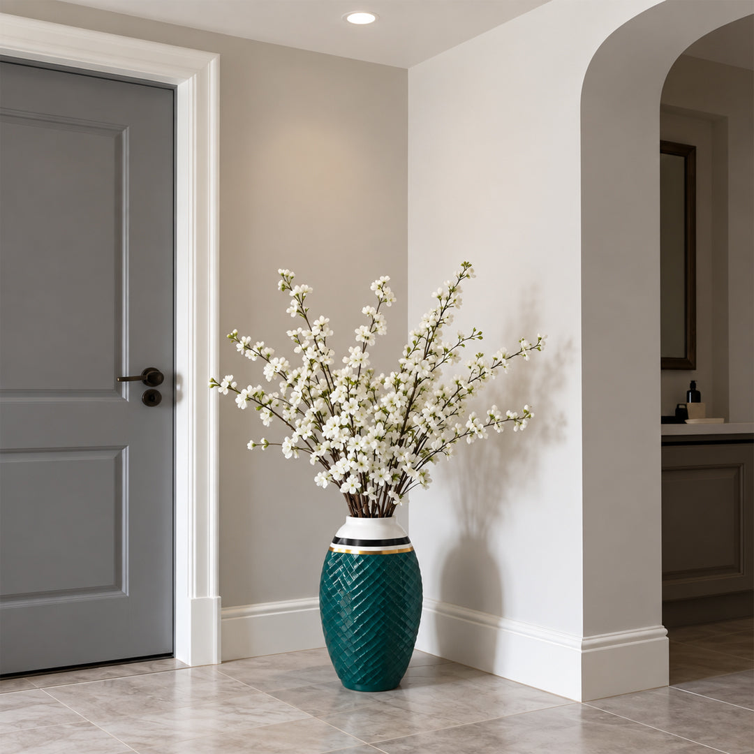 A tall decorative ceramic vase with a textured teal-green body, white band, and gold accent ring, holding white blossoms, placed in a bright entryway corner.