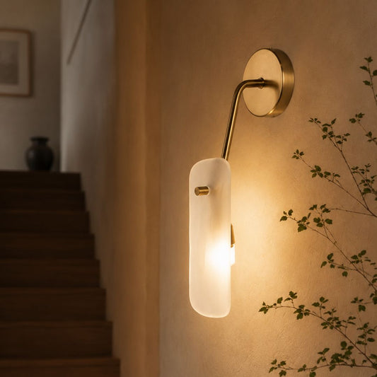 A modern wall sconce light fixture with a vertical frosted glass shade and a curved brass arm, mounted on a beige wall next to a staircase.