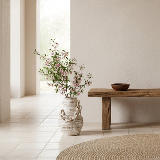 Large chain vase with blooming pink flowers styled beside a wooden bench in a neutral-toned interior setting.