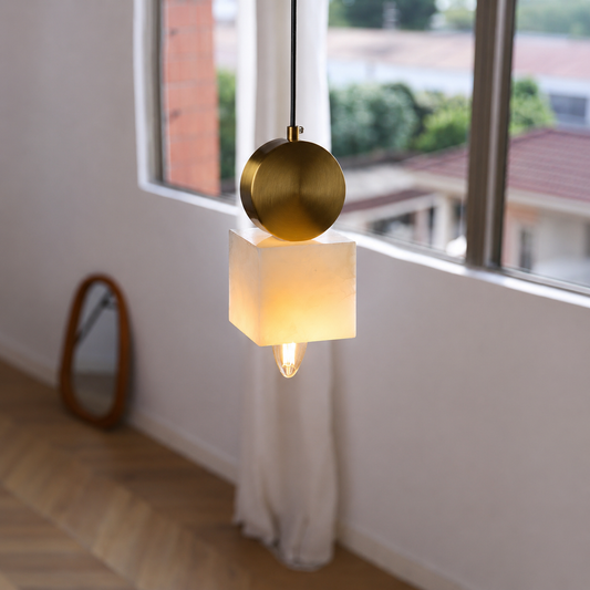 A modern pendant light with a square white fabric shade and a round brushed brass metal cap, hanging in a bright room with a window and light wood floor.